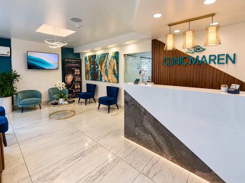 Mostrador de recepción blanco con frente de mármol gris, pared de listones de madera con el logotipo CLINIC MAREN y tres lámparas colgantes doradas.