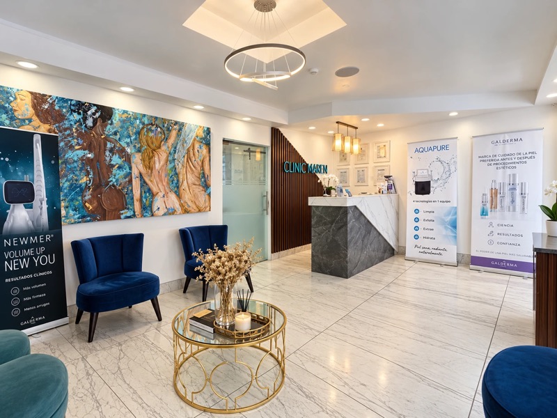 Sala de espera con una pintura de gran formato en tonos azules y dorados, sillones azules, mesa de centro dorada y mostrador de recepción al fondo.