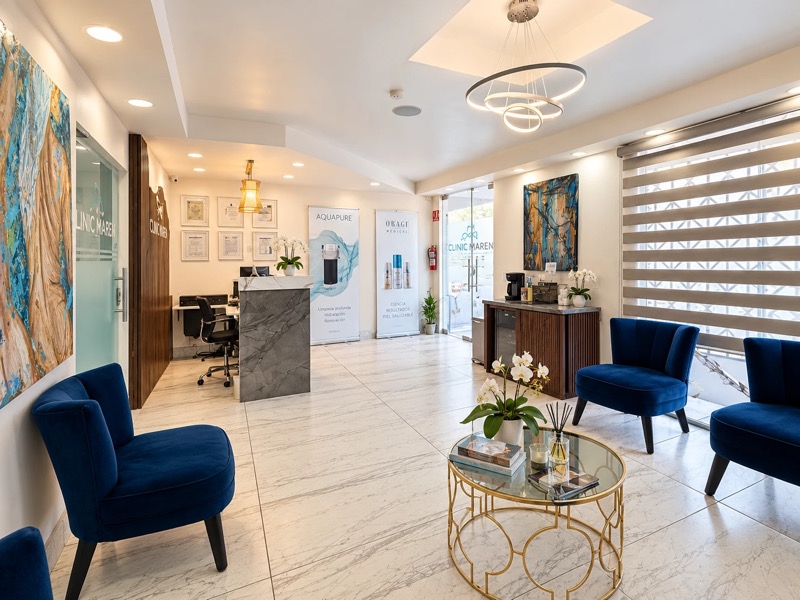 Sala de espera con sillones de terciopelo azul, mesa de centro dorada con orquídeas, estación de café y puerta de cristal de acceso al fondo.